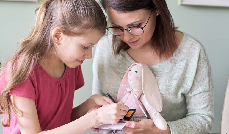Mother and daughter child play together at home, girl and mom make make-up paints cheeks to toy handmade doll bunny sewn on sewing machine
